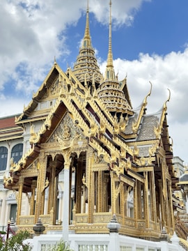 An ornate, gold-clad temple structure with intricate carvings and spires extends upward into a partly cloudy sky. The architecture exhibits elaborate detail and traditional Southeast Asian style.