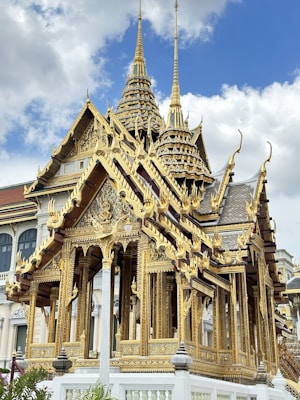 An ornate, gold-clad temple structure with intricate carvings and spires extends upward into a partly cloudy sky. The architecture exhibits elaborate detail and traditional Southeast Asian style.