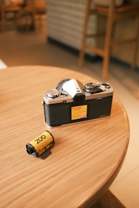 A vintage-style camera with a black and silver body placed on a wooden table next to a roll of Kodak film with a yellow label. The background is softly blurred, hinting at a warm and cozy indoor setting.