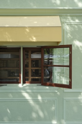 A light green wall features an open wooden window with glass panes, next to a closed window. Above the windows, a yellow awning provides shade. Sunlight filters through tree branches, casting dappled shadows on the wall.
