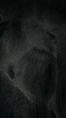 Dark, textured surface with subtle variations in shading, possibly suggesting a soft material or fabric.