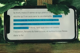 A mobile phone displays a passage from the Bible, specifically Galatians 6. The text is written in French. Some sentences are highlighted in blue indicating their importance. The phone is placed on a wooden surface with green leaves around it, suggesting an outdoor setting.