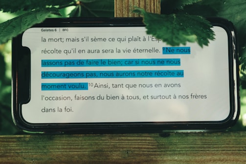 A mobile phone displays a passage from the Bible, specifically Galatians 6. The text is written in French. Some sentences are highlighted in blue indicating their importance. The phone is placed on a wooden surface with green leaves around it, suggesting an outdoor setting.