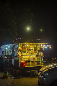 A brightly lit mobile street food stall at night, surrounded by various packaged snacks and beverages. A vendor stands nearby, serving a customer. The area is dimly lit except for the stall, creating a contrast against the dark surroundings.