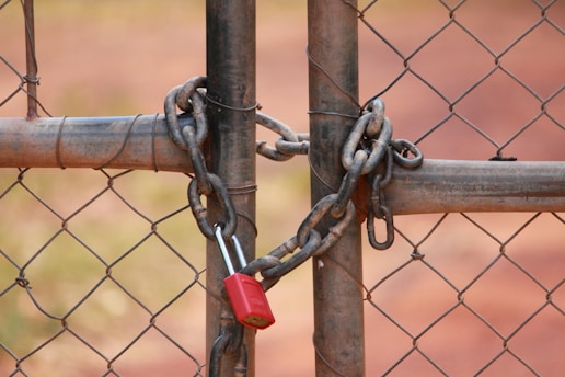 A metal chain-link fence is secured by a heavy chain and a red padlock. The focus is on the closed lock and intertwined chain, signifying security or restriction. The background is out of focus, with reddish-brown earth tones.