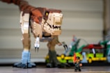 A large Lego dinosaur, resembling a Tyrannosaurus rex, is positioned prominently on a surface. In front of it stands a small Lego figure of a person, holding a red item in one hand. The background includes a green Lego car and additional Lego structures, partially blurred.