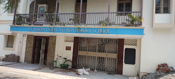 A building facade with a blue sign reading 'St. Joseph of Cluny Montessori School.' The balcony features potted plants and the entrance includes two metal gates. A green bicycle is parked near the entrance, with bricks and construction materials stacked on the side.