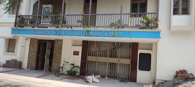 A building facade with a blue sign reading 'St. Joseph of Cluny Montessori School.' The balcony features potted plants and the entrance includes two metal gates. A green bicycle is parked near the entrance, with bricks and construction materials stacked on the side.