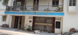 A building facade with a blue sign reading 'St. Joseph of Cluny Montessori School.' The balcony features potted plants and the entrance includes two metal gates. A green bicycle is parked near the entrance, with bricks and construction materials stacked on the side.