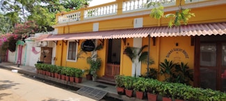 A bright yellow building with decorative white trim houses a guest house. The entrance is adorned with several potted green plants, a red wooden gate, and a black sign. The facade features a red sloped awning. Trees and pink flowering plants partially shade the scene.