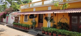A bright yellow building with decorative white trim houses a guest house. The entrance is adorned with several potted green plants, a red wooden gate, and a black sign. The facade features a red sloped awning. Trees and pink flowering plants partially shade the scene.