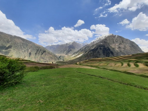 A vast, lush green landscape stretches across the foreground with terraced fields leading up to majestic snow-capped mountains under a bright, partly cloudy sky.