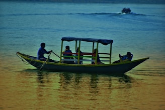 A small boat with a canopy floats on the water with several people seated inside. The scene captures a man rowing while others relax and take photos. The water reflects the colors of the sky, creating a serene and peaceful atmosphere.