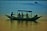 A small boat with a canopy floats on the water with several people seated inside. The scene captures a man rowing while others relax and take photos. The water reflects the colors of the sky, creating a serene and peaceful atmosphere.