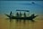 A small boat with a canopy floats on the water with several people seated inside. The scene captures a man rowing while others relax and take photos. The water reflects the colors of the sky, creating a serene and peaceful atmosphere.