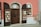 A storefront with a glass door and two display cabinets filled with jewelry and decorative items. The building has a warm pink and cream exterior with a brown door and shutters. The displays feature various jewelry pieces, likely made of crystal or glass, placed on white stands.