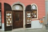 A storefront with a glass door and two display cabinets filled with jewelry and decorative items. The building has a warm pink and cream exterior with a brown door and shutters. The displays feature various jewelry pieces, likely made of crystal or glass, placed on white stands.