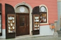 A storefront with a glass door and two display cabinets filled with jewelry and decorative items. The building has a warm pink and cream exterior with a brown door and shutters. The displays feature various jewelry pieces, likely made of crystal or glass, placed on white stands.