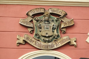A decorative crest or emblem mounted on a wall, featuring ornate designs and inscriptions in German. The emblem includes a depiction of a castle or building set within a shield and surrounded by intricate scrollwork. The words 'Stadt-Apotheke' and '1620' are prominently inscribed.