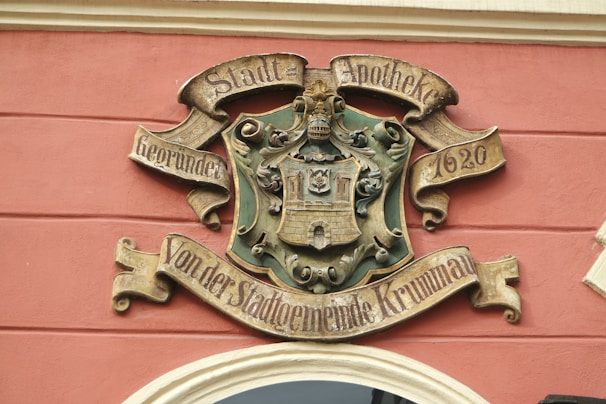 A decorative crest or emblem mounted on a wall, featuring ornate designs and inscriptions in German. The emblem includes a depiction of a castle or building set within a shield and surrounded by intricate scrollwork. The words 'Stadt-Apotheke' and '1620' are prominently inscribed.