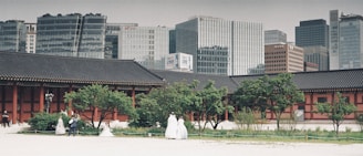 A traditional Korean palace courtyard is set against a backdrop of modern skyscrapers. The courtyard features a large Korean-style building with tiled roofs and columns, surrounded by greenery. Several people dressed in traditional Korean hanboks are visible among the trees, suggesting a cultural or historical theme. The contrast between the historical architecture and the modern urban skyline highlights a blend of old and new.