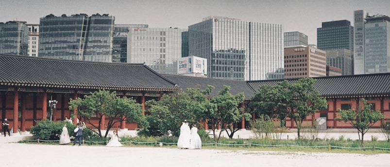 A traditional Korean palace courtyard is set against a backdrop of modern skyscrapers. The courtyard features a large Korean-style building with tiled roofs and columns, surrounded by greenery. Several people dressed in traditional Korean hanboks are visible among the trees, suggesting a cultural or historical theme. The contrast between the historical architecture and the modern urban skyline highlights a blend of old and new.