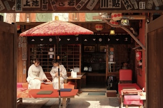 Two people dressed in traditional Japanese kimonos are sitting on red benches under a red and white traditional umbrella in front of a wooden structure adorned with Japanese decorations and paper lanterns.