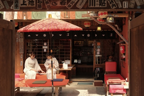 Two people dressed in traditional Japanese kimonos are sitting on red benches under a red and white traditional umbrella in front of a wooden structure adorned with Japanese decorations and paper lanterns.