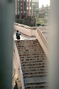 A person wearing a school uniform and carrying a backpack ascends a weathered staircase on an urban pedestrian bridge. The scene is set in a cityscape with a tall modern building and some greenery in the background.