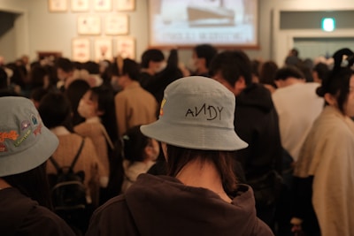 A crowded room filled with people who are closely gathered together. In the foreground, a person wearing a light blue bucket hat with the name 'ANDY' printed on it is visible. The atmosphere appears busy, as many individuals are facing towards a screen or display at the far end of the room.