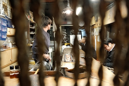A cozy, dimly lit restaurant interior with wooden furniture and various objects scattered around. Several people are seated at a bar or counter, with one person standing and appearing to engage with others. Decorative elements and a collection of bottles line the shelves, while warm lighting creates an intimate atmosphere.