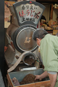 A person in a green garment is scooping tea leaves from a container attached to a traditional tea roasting machine. The machine has Japanese characters on it, and there's a busy store with various items in the background.