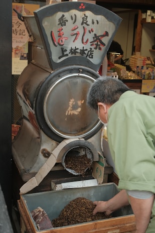 A person in a green garment is scooping tea leaves from a container attached to a traditional tea roasting machine. The machine has Japanese characters on it, and there's a busy store with various items in the background.