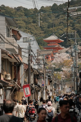 A bustling street scene with numerous people walking, surrounded by traditional Japanese architecture. A prominent red pagoda can be seen in the distance, partially obscured by trees. The street is lively with a mix of tourists and locals, and cherry blossoms are visible in the background, indicating springtime.