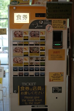 A vending machine for purchasing food tickets, featuring a user interface with photos of various meal options. Prominent instructions in both Japanese and English guide users on how to obtain and use the tickets. The machine accepts coins and notes, and different food selections are available, such as pizza and other dishes.