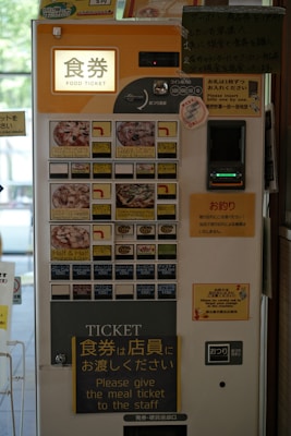 A vending machine for purchasing food tickets, featuring a user interface with photos of various meal options. Prominent instructions in both Japanese and English guide users on how to obtain and use the tickets. The machine accepts coins and notes, and different food selections are available, such as pizza and other dishes.