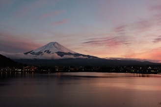 A stunning view of a snow-capped mountain, likely Mount Fuji, during sunset. The sky is painted with hues of pink, orange, and purple, while the mountain stands majestically in the background. In the foreground, a calm body of water reflects the serene colors of the sky.