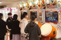 Several people are engaged with arcade games, focusing on rhythm games with large drum controllers. The players appear focused and are wearing masks, indicating a casual, indoor setting in an arcade filled with vibrant colors.