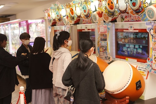 Several people are engaged with arcade games, focusing on rhythm games with large drum controllers. The players appear focused and are wearing masks, indicating a casual, indoor setting in an arcade filled with vibrant colors.