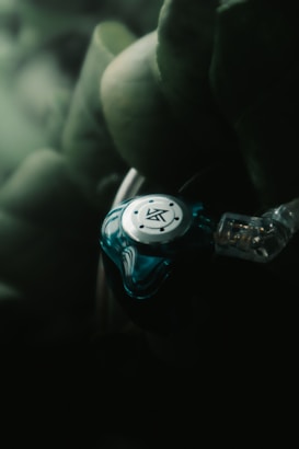 A close-up view of a blue and silver earphone with intricate design, surrounded by soft-focus greenery. The earphone appears sleek and modern, with visible wiring.
