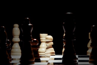 A chessboard with several chess pieces arranged in strategic positions. The focus is on a white pawn, illuminated by a dim light, creating contrasts with the darker pieces around it. The scene is set against a dark background, enhancing the dramatic effect.