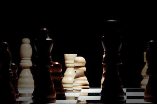 A chessboard with several chess pieces arranged in strategic positions. The focus is on a white pawn, illuminated by a dim light, creating contrasts with the darker pieces around it. The scene is set against a dark background, enhancing the dramatic effect.