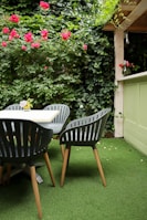 An outdoor patio area features four dark green chairs with wooden legs positioned around a light-colored table. The ground is covered with artificial grass, and the backdrop consists of lush green ivy vines and red roses climbing a wooden trellis. A vase with flowers sits on a ledge nearby, adding a decorative touch to the space.