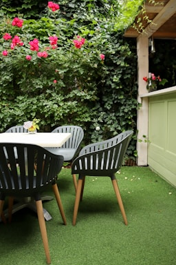 An outdoor patio area features four dark green chairs with wooden legs positioned around a light-colored table. The ground is covered with artificial grass, and the backdrop consists of lush green ivy vines and red roses climbing a wooden trellis. A vase with flowers sits on a ledge nearby, adding a decorative touch to the space.