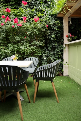 An outdoor patio area features four dark green chairs with wooden legs positioned around a light-colored table. The ground is covered with artificial grass, and the backdrop consists of lush green ivy vines and red roses climbing a wooden trellis. A vase with flowers sits on a ledge nearby, adding a decorative touch to the space.