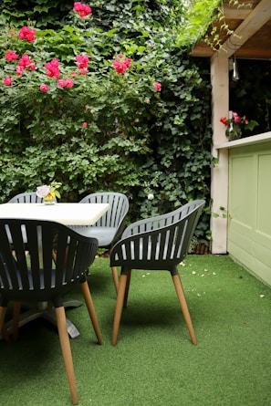 An outdoor patio area features four dark green chairs with wooden legs positioned around a light-colored table. The ground is covered with artificial grass, and the backdrop consists of lush green ivy vines and red roses climbing a wooden trellis. A vase with flowers sits on a ledge nearby, adding a decorative touch to the space.