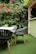 An outdoor patio area features four dark green chairs with wooden legs positioned around a light-colored table. The ground is covered with artificial grass, and the backdrop consists of lush green ivy vines and red roses climbing a wooden trellis. A vase with flowers sits on a ledge nearby, adding a decorative touch to the space.