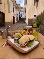 A wooden table set in an outdoor alleyway features a sandwich with lettuce and tomato, accompanied by a portion of fries, condiments, and a drink served in a clear glass with ice. A bottle of Inca Kola is also visible. The background shows a narrow, cobbled street with traditional stone buildings, and a clear blue sky is overhead.