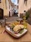 A wooden table set in an outdoor alleyway features a sandwich with lettuce and tomato, accompanied by a portion of fries, condiments, and a drink served in a clear glass with ice. A bottle of Inca Kola is also visible. The background shows a narrow, cobbled street with traditional stone buildings, and a clear blue sky is overhead.