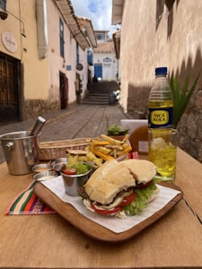 A wooden table set in an outdoor alleyway features a sandwich with lettuce and tomato, accompanied by a portion of fries, condiments, and a drink served in a clear glass with ice. A bottle of Inca Kola is also visible. The background shows a narrow, cobbled street with traditional stone buildings, and a clear blue sky is overhead.